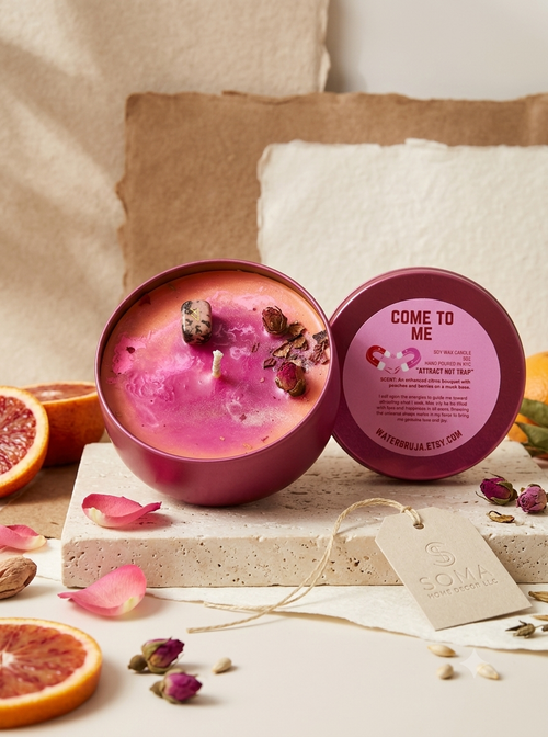 An open round candle tin and its matching lid are displayed on a stone slab with citrus slices, dried rose petals, and a paper tag. The candle wax is bright pink, and the lid label reads 'COME TO ME' with 'MATERNALLYA KTYE.