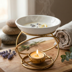 A white ceramic candle warmer bowl with a gold metal stand, shown lit with a small tealight candle inside. The setup is styled on a wooden surface with lavender sprigs, a rolled towel, and stacked stones in the background.