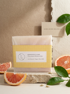 A packaged bar of soap labeled 'GRAPEFRUIT SOAP Cleansing & Moisturizing' sits on a stone slab with grapefruit slices and green leaves around it. A small card reading 'SOMA HOME DEC LLC' is behind the soap against a textured beige wall.