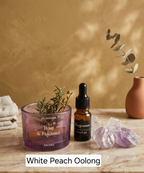 A styled tabletop scene showing a purple glass cold air diffuser candle and a small amber dropper bottle labeled 'Fragrance Fresh air' beside purple crystals, with a warm textured tan wall and eucalyptus in a terracotta vase in the background. Text at the bottom reads 'White Peach Oolong'.