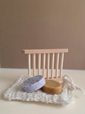 Two round handmade soap bars, one lavender with dark specks and one tan, displayed on a white crochet soap dish in front of a small wooden soap dish holder against a neutral beige background.
