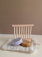 Two round handmade soap bars, one lavender with dark specks and one tan, displayed on a white crochet soap dish in front of a small wooden soap dish holder against a neutral beige background.