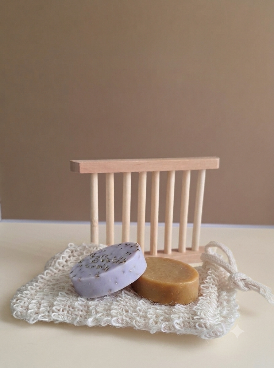 Two round handmade soap bars, one lavender with dark specks and one tan, displayed on a white crochet soap dish in front of a small wooden soap dish holder against a neutral beige background.
