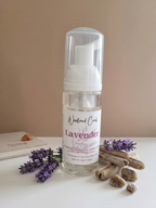 A bottle of Woodland Creek Lavender Hand Sanitizer with a clear cap and white pump, styled on a white surface with lavender sprigs, driftwood, and stones against a beige background.