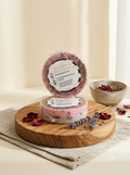 Two round bars of soap, one standing upright and one lying flat, displayed on a round wooden board with dried rose petals and lavender sprigs. A small bowl of dried petals sits in the background.