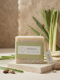 A wrapped bar of lemonade soap sits on a stone slab with lemongrass stems and small lemongrass bulbs around it. The soap is wrapped in a pale green paper band with a white label that reads 'LEMONADE SOAP' and 'SOMA' on the hanging tag.