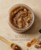 A clear round jar filled with a coarse brown scrub or mask sits on a beige stone-like surface. A wooden spoon with amber-brown product and a glass dropper with honey-like amber liquid are placed nearby, along with small brown sugar cubes.
