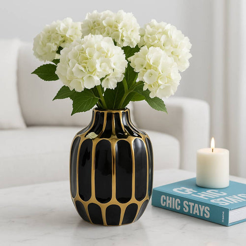 Black and gold vase with white flowers on a table next to a book and candle.