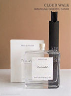 A clear glass reed diffuser bottle labeled 'Fascinated.' stands in front of a white branded box and a tall black reed holder, with the text 'CLOUD WALK' and 'AURA RELAX / COMFORT / NATURE' above on a warm beige background.