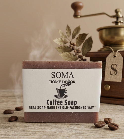 A rectangular bar of SOMA Home Decor Coffee Soap stands on a rustic wooden surface with scattered coffee beans. The soap has a white label with black text and a coffee cup illustration, with a warm brown soap base visible at the top and bottom.