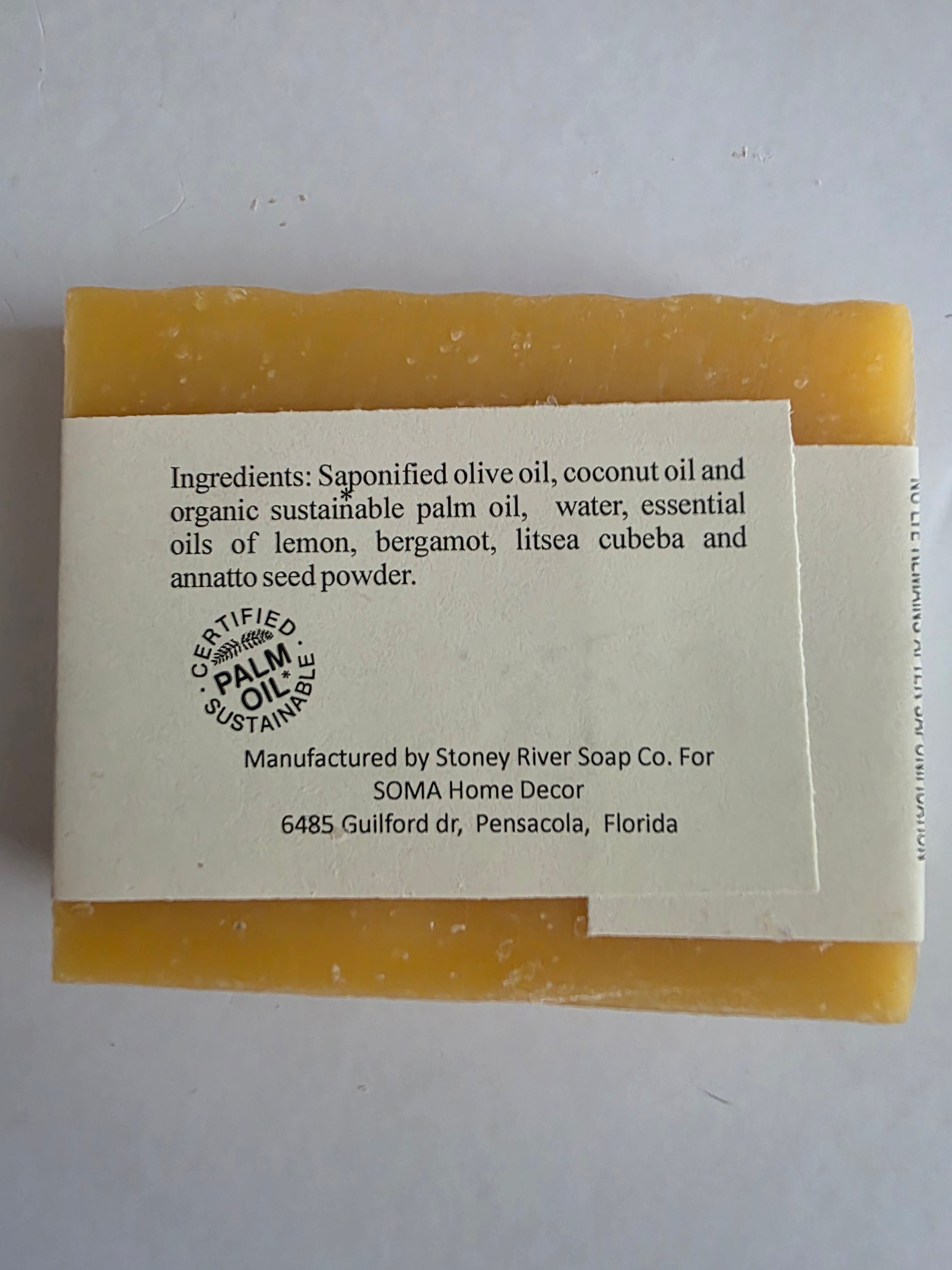 A rectangular bar of handmade soap in a warm yellow-orange color, wrapped with a white paper label showing the brand 'Storey River Soap Co.' and other product details.
