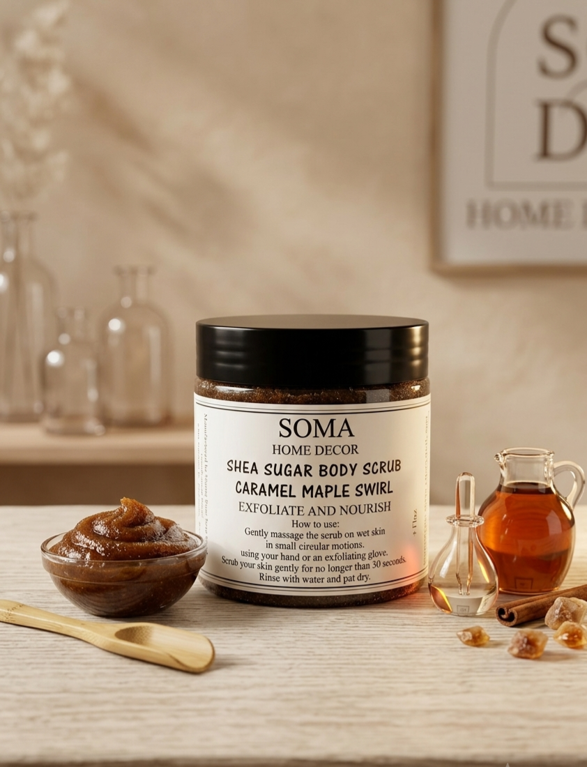 A jar of Soma Home Decor Shea Sugar Body Scrub with a black lid and white label, styled on a wooden surface with a small bowl of scrub, a wooden spoon, a glass bottle of amber liquid, and cinnamon sticks against a warm home decor background.