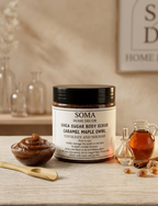 A jar of Soma Home Decor Shea Sugar Body Scrub with a black lid and white label, styled on a wooden surface with a small bowl of scrub, a wooden spoon, a glass bottle of amber liquid, and cinnamon sticks against a warm home decor background.